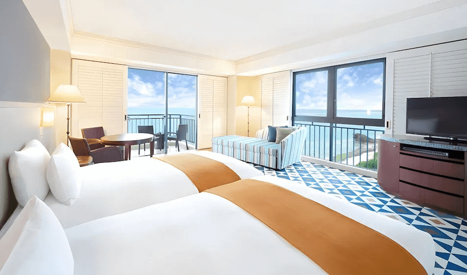 【讀谷村住宿】"讀古村住宿精選示意圖"「沖繩阿利比拉日航度假飯店的海景雙床房，房內採用藍白色調搭配橘色床巾，大片落地窗可直接眺望湛藍海景，營造明亮度假的舒適氛圍。」