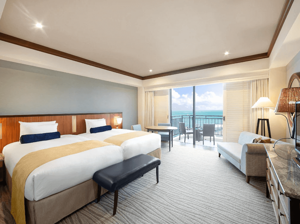 【讀谷村住宿】「沖繩阿利比拉日航度假飯店的寬敞雙床客房，採用溫潤木質與米色調設計，落地窗外即是海景陽台，讓房內充滿自然光線與療癒海景。」