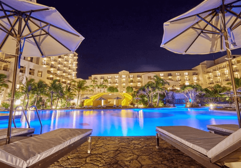 【讀谷村住宿】「沖繩阿利比拉日航度假飯店的夜間泳池景色，燈光映照在池水上，周圍熱帶植栽與度假建築營造浪漫且夢幻的度假夜晚。」