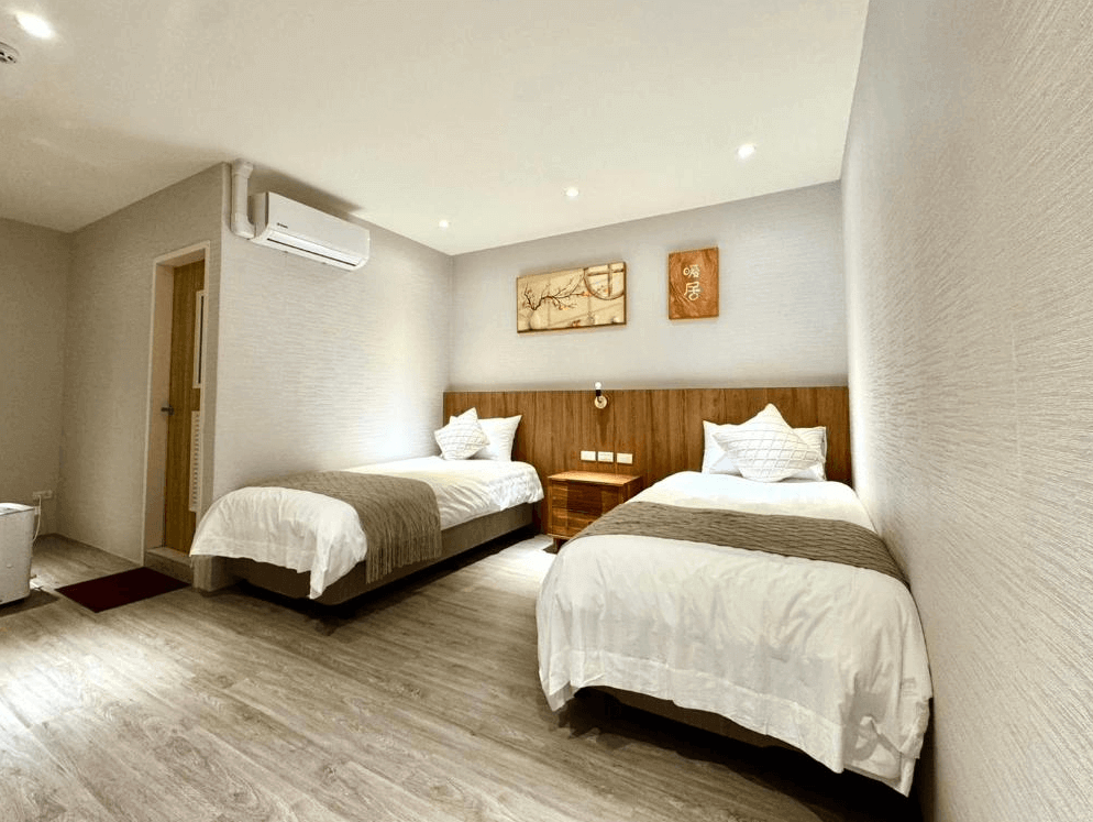 【大溪住宿】湧山水民宿 標準雙床房，淺木色調空間簡約舒適，設有冷氣與暖色照明，床頭掛有書法與木刻裝飾，整體乾淨雅緻。
