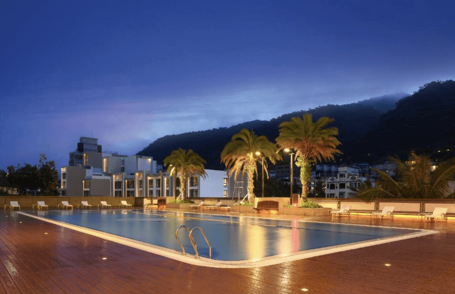 【宜蘭溫泉飯店】「礁溪長榮鳳凰酒店戶外泳池夜景，泳池周圍種植棕櫚樹並設有休憩躺椅，背景為山景與飯店建築，在夜間燈光映照下充滿度假氛圍。」