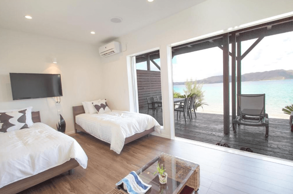 【奄美大島住宿】「美阿丹海灘飯店客房景觀：房內兩張單人床、大片落地窗直面碧藍海景與木質露台，房間明亮乾淨、空間舒適。」