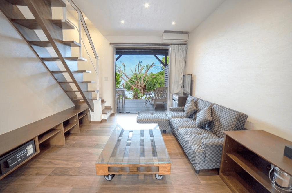 【奄美大島住宿】「美阿丹海灘飯店樓中樓客房：一樓客廳設計含沙發與茶几，通往室外露台可欣賞熱帶植物與大海，室內採木質風格溫馨舒適。」