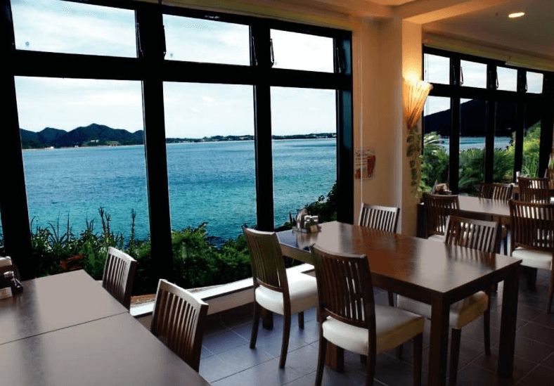 【奄美大島住宿】「美阿丹海灘飯店餐廳景觀：用餐區面向清澈海景與山脈，全景落地窗提供壯麗海岸線視野，座位舒適明亮。」