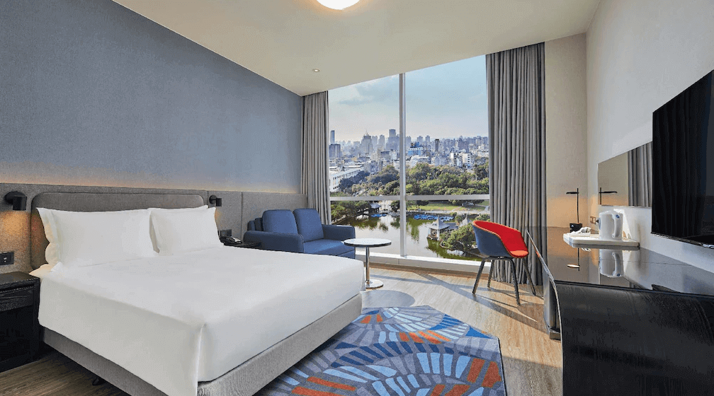 【台中火車站住宿】"台中火車站住宿精選示意圖"「臺中公園智選假日飯店客房，大片落地窗可眺望台中市區與綠意景觀，房內配置大床、藍色雙人沙發、書桌與現代風格地毯。」