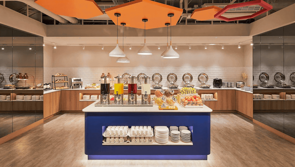 【台中火車站住宿】「臺中公園智選假日飯店自助式早餐餐廳，中央島台擺放果汁、麥片與水果，四周陳列中西式早餐料理，空間明亮寬敞。」