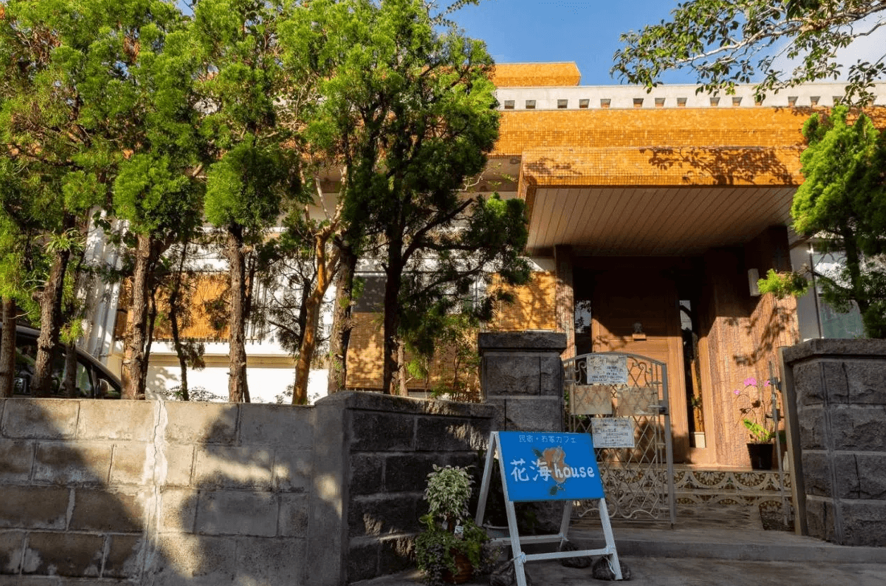 【奄美大島住宿】「花海house（Kami House）外觀：被綠樹環繞的民宿建築，以溫暖的褐色外牆與藍色指示牌為特色，入口處明亮整潔。」