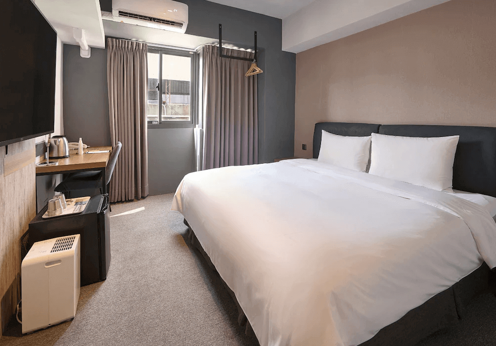 【台中火車站住宿】「逗點 inn Hotel 的標準雙人房，採用柔和灰色系與簡約木質設計，搭配大張雙人床、書桌與迷你冰箱，房間明亮舒適、風格簡潔。」