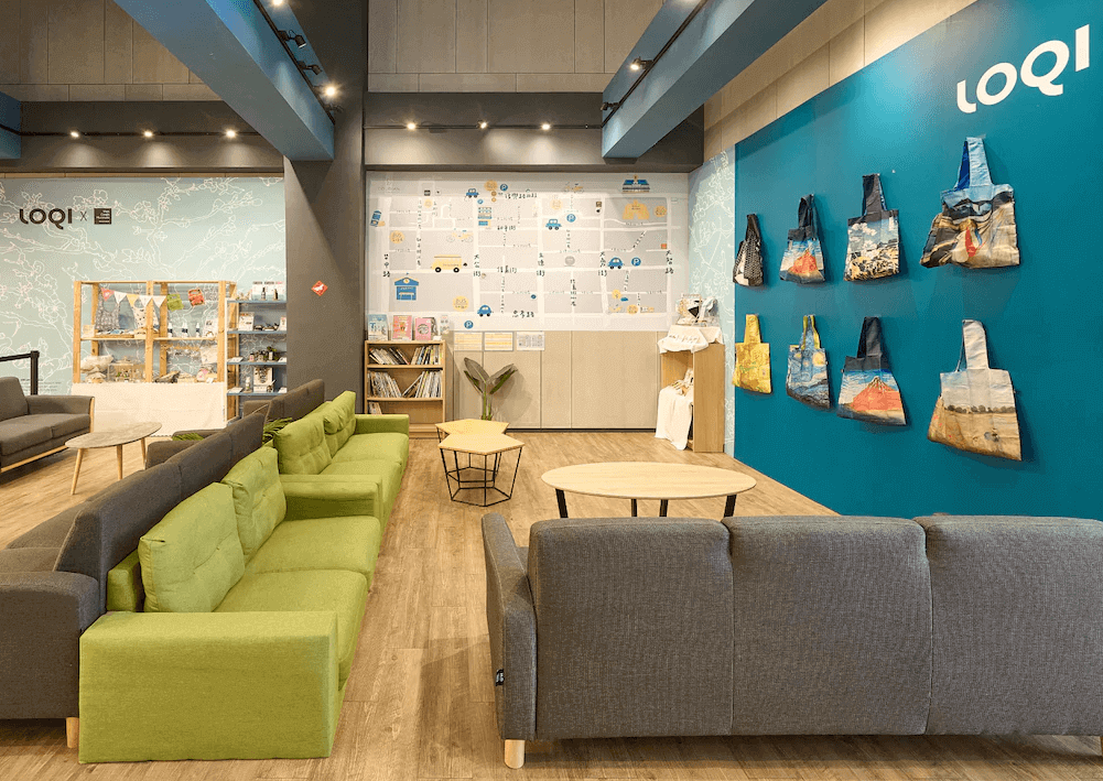 【台中火車站住宿】「逗點 inn Hotel 的公共交誼廳，設有多組綠灰色沙發、木質桌椅與大型地圖裝飾牆，空間溫馨活潑，並展示多款 LOQI 設計提袋。」