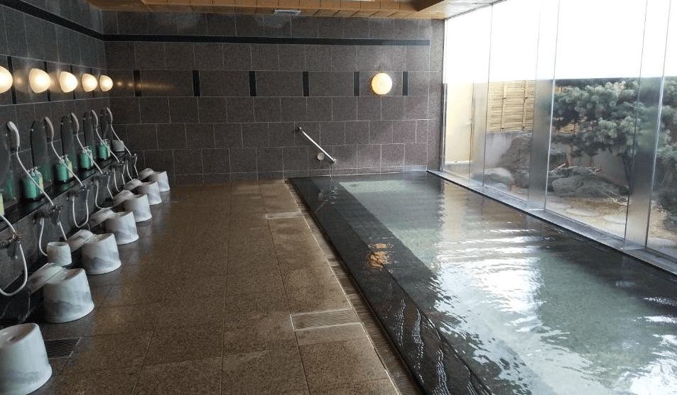 【北見住宿】露櫻酒店北見大通西店的大浴場，設有整排淋浴座與溫泉池，牆面以深色石材打造，大片玻璃窗外可見庭園景致。