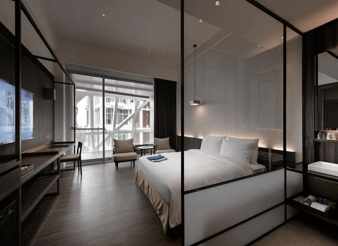 【宜蘭溫泉飯店】「風華渡假酒店・礁溪館客房內景，現代極簡風格設計，大片玻璃隔間連接浴室區域，房內配備大床、書桌、休憩椅與落地窗，採光通透明亮。」