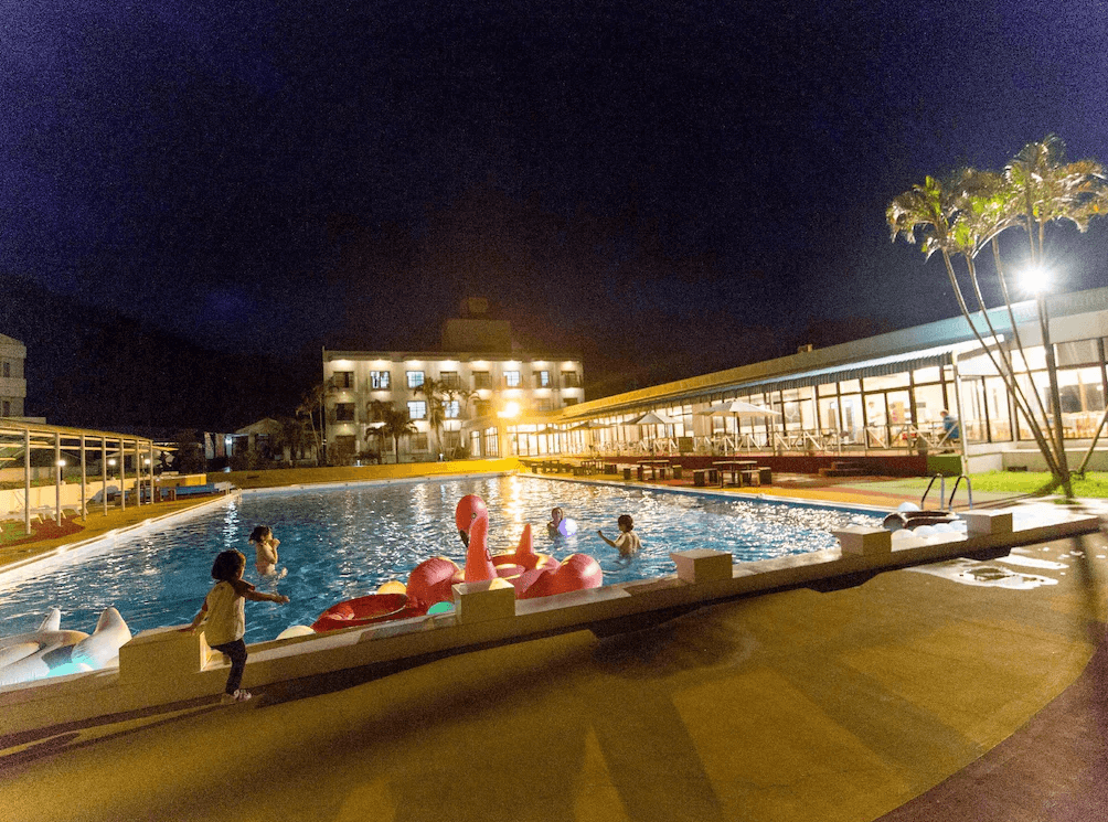 【奄美大島住宿】「龍美 Caretta 飯店（Hotel Caretta Amami）夜間戶外泳池，孩子與旅客在池中戲水，飯店建築倒映在水面上，呈現熱鬧度假氛圍。」