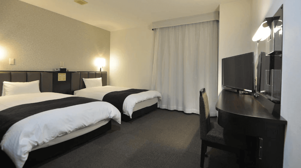 【霧島住宿】APA 飯店的雙床客房：寬敞的 APA 飯店雙床客房，內有兩張單人床、一張靠牆的長桌、電視和椅子，窗邊掛有白色窗簾。