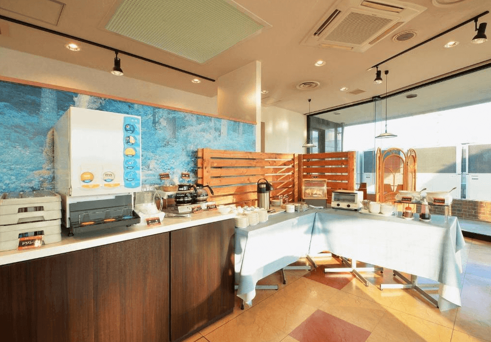 【霧島住宿】APA 飯店的自助早餐區：APA 飯店內提供自助式早餐的區域，設有飲料機、咖啡機、烤麵包機，以及擺放各式小菜和餐具的長桌，空間明亮。
