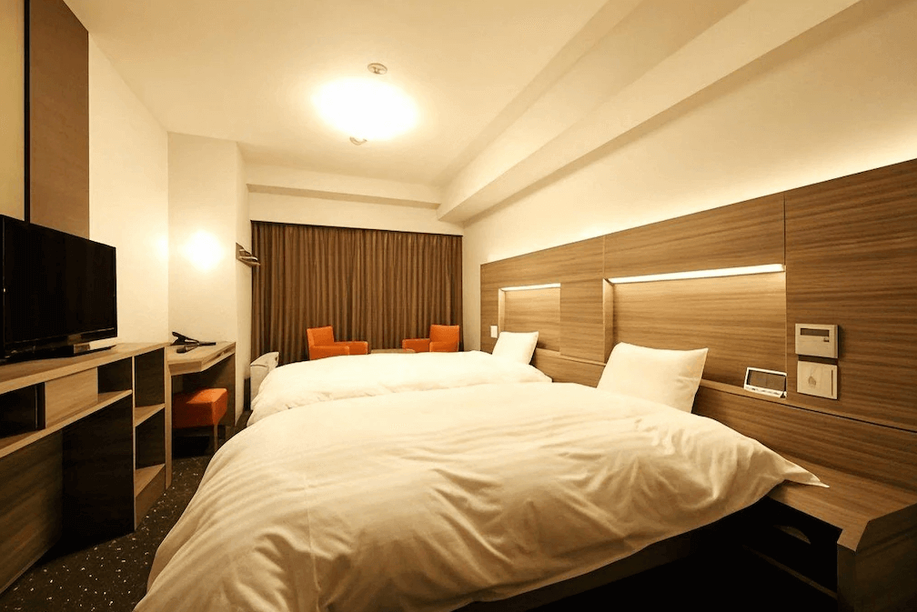 【鹿兒島住宿】Dormy Inn飯店-鹿兒島天然溫泉 的舒適雙床客房，採用溫暖的木質調裝潢，床頭牆面設計有柔和的間接照明，靠窗處配置兩張亮眼的橘色休閒椅，提供旅客放鬆休憩的空間。