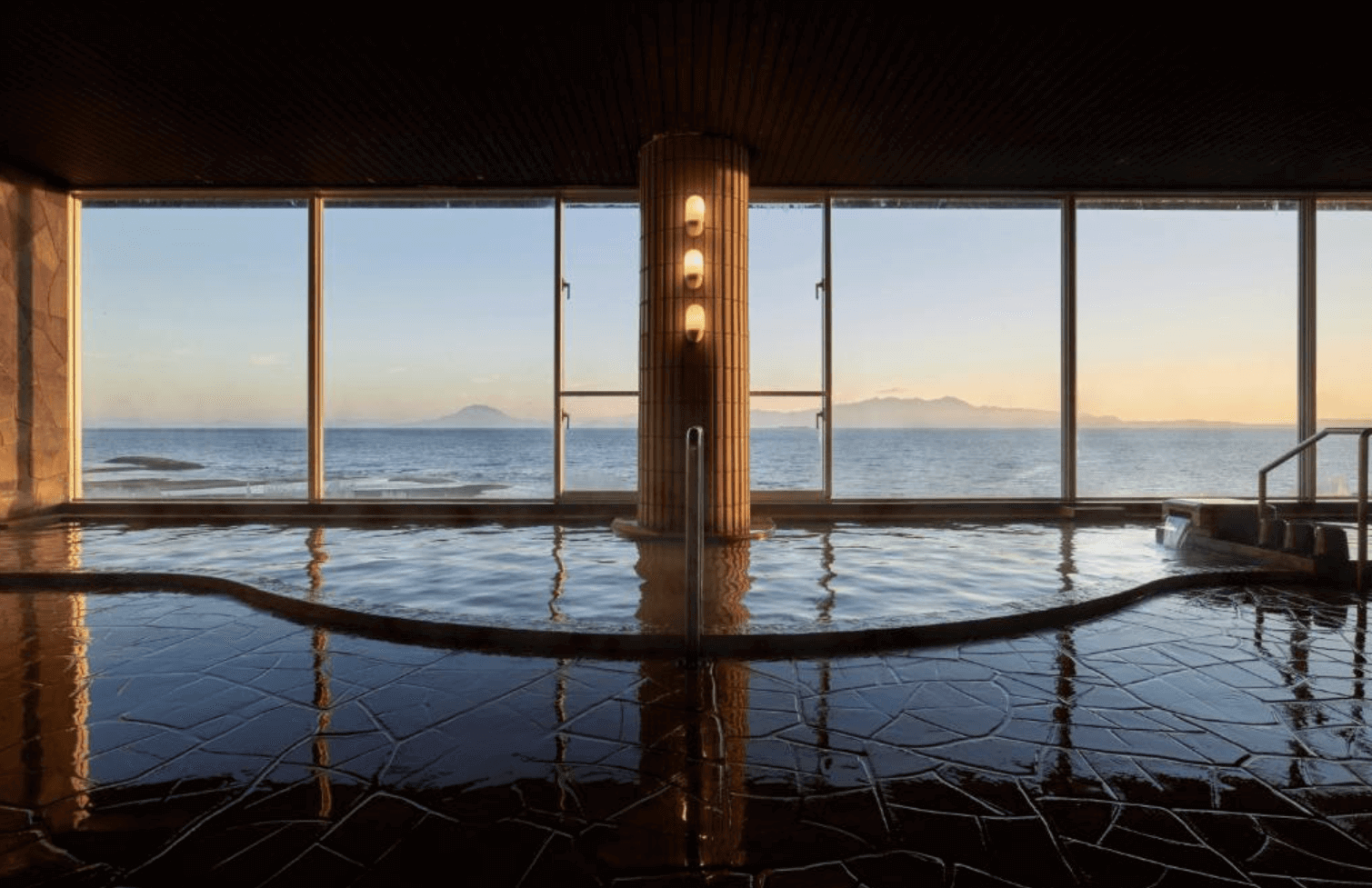 【指宿住宿】Koran之湯錦江樓 (Ryokan Koran no Yu Kinkouroh) 的天然溫泉大浴場，以整面落地玻璃窗打造無遮蔽視野，讓旅客邊泡湯邊欣賞遠方櫻島火山與平靜海面的絕景。