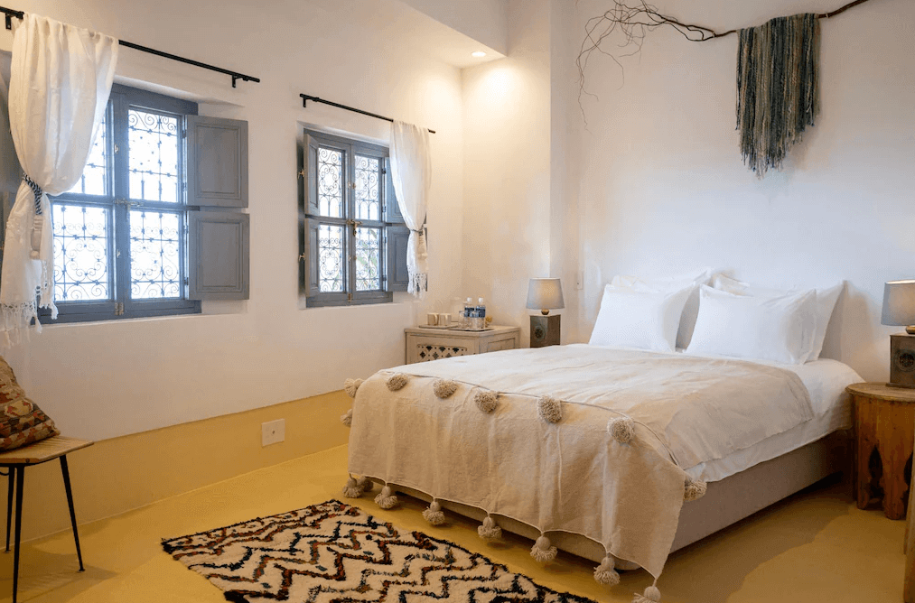 【沖繩南城住宿】Riad Lamp 的摩洛哥風情客房，以純白牆面搭配帶有精緻鐵窗花的藍色木窗，床上鋪設帶有絨球裝飾的異國風織品，營造出彷彿置身北非的獨特住宿體驗。