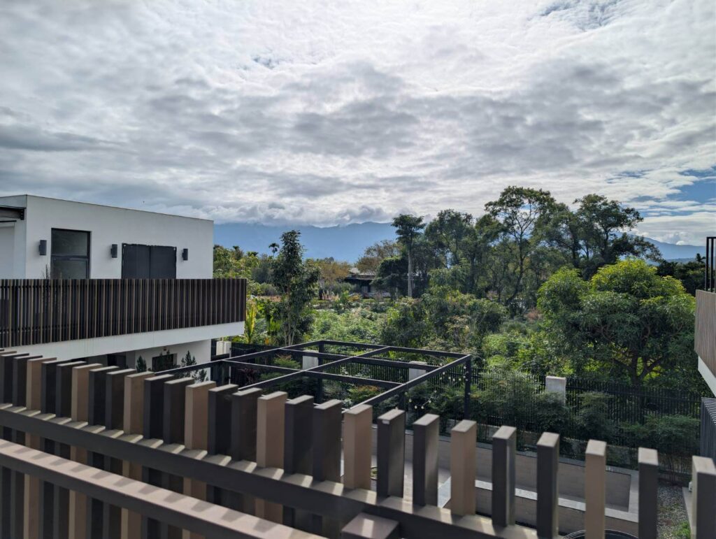 【元湯溫泉居住宿】「花蓮《元湯溫泉居》二樓陽台景色：白色現代風建築前方，是被綠意包圍的庭園與大片樹林，遠方可見層疊山景與雲海，在陽光穿透雲層下呈現清新而靜謐的度假氛圍。」