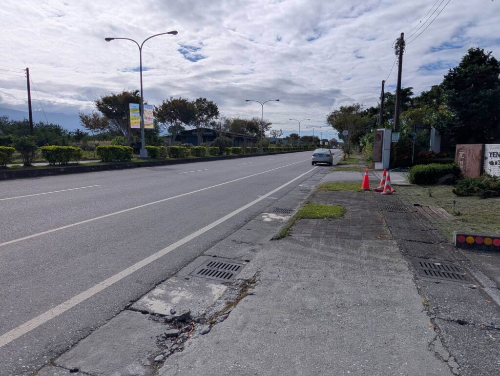〈元湯溫泉居〉外部道路景觀，顯示筆直公路、行道樹與旅宿入口處指示牌，可見停放車輛與交通錐，呈現住宿周邊環境的寧靜與開闊感。