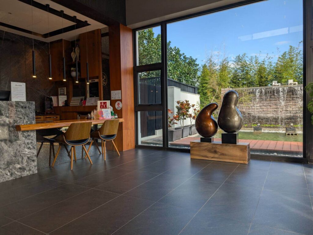 「元湯溫泉居大廳空間，落地窗外可看到水池與瀑布景觀，室內擺放兩座造型藝術雕塑，旁側為櫃檯及木質桌椅組，呈現溫暖舒適的迎賓氛圍。」