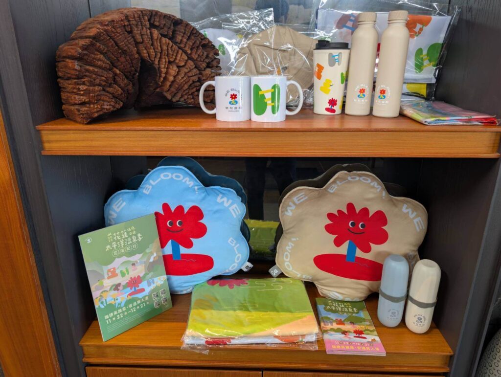 「元湯溫泉居大廳展售區，展示太平洋溫泉季周邊商品，包括可愛花朵造型抱枕、馬克杯、環保瓶、毛巾與活動DM等彩色設計商品。」