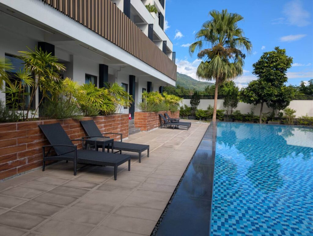 「元湯溫泉居泳池旁的池畔躺椅區，搭配棕色植栽牆、棕櫚樹與藍色泳池，展現舒適的南國度假風景。」