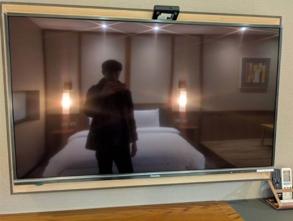 〈元湯溫泉居〉房間內的 Panasonic 大尺寸電視，螢幕反射出溫暖燈光下的床鋪與室內擺設。