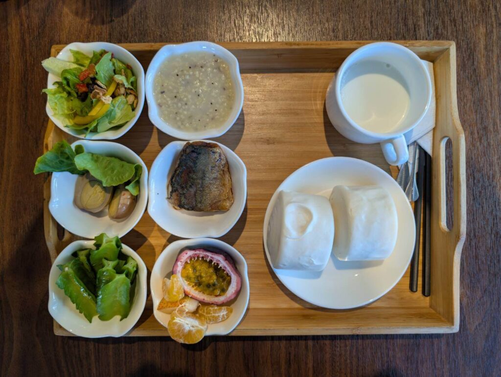 〈元湯溫泉居〉早餐套餐，包含生菜沙拉、粥品、滷蛋、烤魚、鮮蔬、百香果與橘子，以及搭配兩顆饅頭與一杯熱牛奶的中式早餐組合。