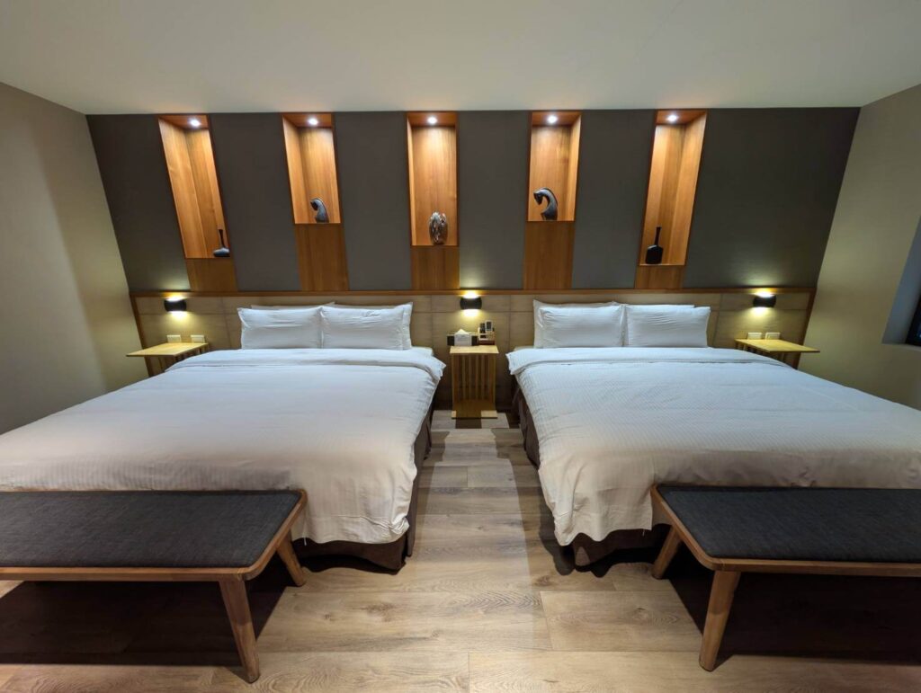 〈元湯溫泉居〉洋式四人房內景，兩張大床整齊排列，背牆以木質展示壁龕與燈光打造溫馨質感。