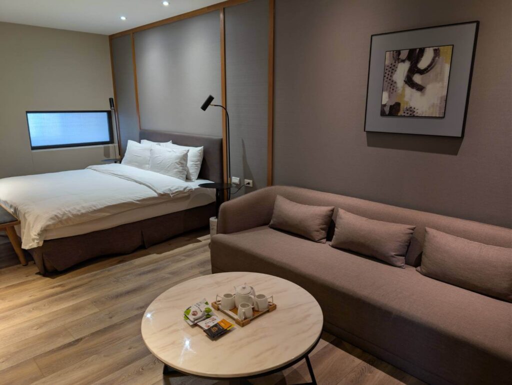 元湯溫泉居的洋式雙人房內部，呈現現代風格。房間設有一張鋪有白色寢具的大床和一張淺棕色三人沙發。沙發前的圓形茶几上擺放著精緻茶具。房間採用木紋地板，牆面以灰色調為主，營造出簡約舒適的住宿環境。