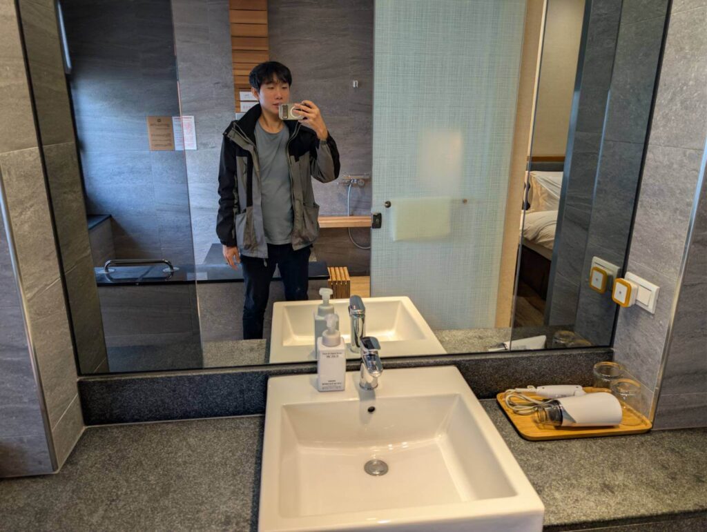 〈元湯溫泉居〉浴室空間，寬大的鏡面映照出洗手台、吹風機托盤與泡湯區的灰石設計風格。