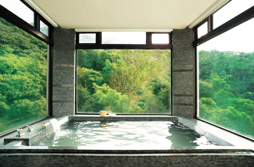 北投大地酒店 (The Gaia Hotel) 的景觀溫泉湯池，以灰黑色石材打造沉穩質感，透過 L 型大面玻璃窗，將戶外濃密的森林綠意盡收眼底，享受被大自然包圍的泡湯體驗。