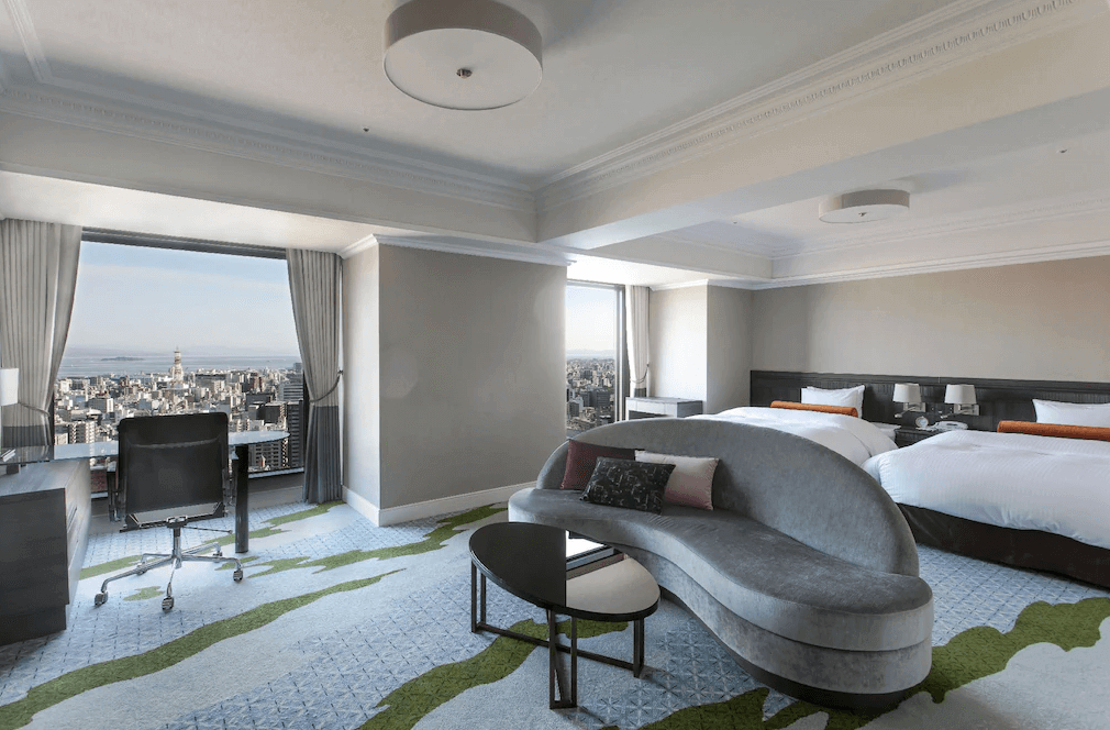 【鹿兒島住宿】城山觀光飯店 (SHIROYAMA HOTEL kagoshima) 的現代風格轉角客房，房內設有兩張單人床及灰色弧形沙發，透過兩面寬敞的觀景窗，可將鹿兒島市區的開闊美景盡收眼底。
