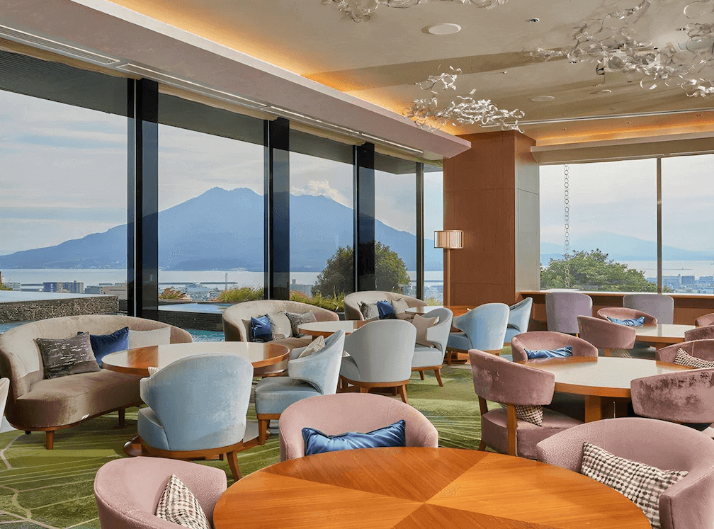 【鹿兒島住宿】城山觀光飯店 (SHIROYAMA HOTEL kagoshima) 的景觀交誼廳與餐廳區域，擁有整面落地窗設計，搭配粉色與藍色的優雅沙發座席，提供旅客欣賞櫻島火山絕景的休憩空間。