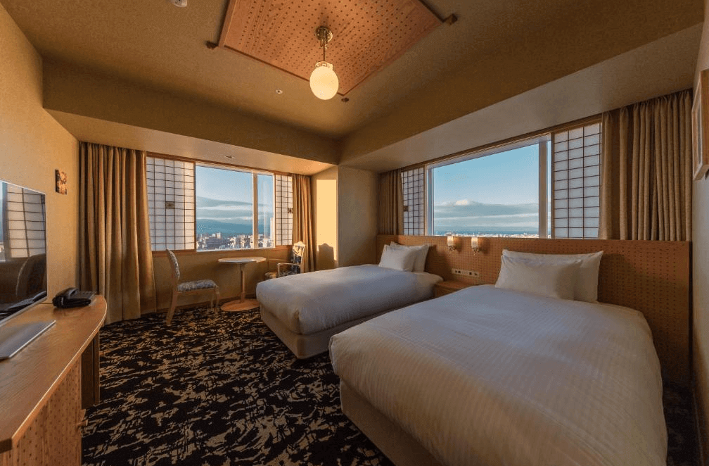 【大分住宿】大分JR九州布魯斯姆酒店 (JR Kyushu Hotel Blossom Oita) 的舒適客房視角,兩張寬敞的單人床鋪設潔白寢具,透過大面觀景窗引入柔和的自然光線與城市天際線,營造出溫暖寧靜的住宿氛圍。
