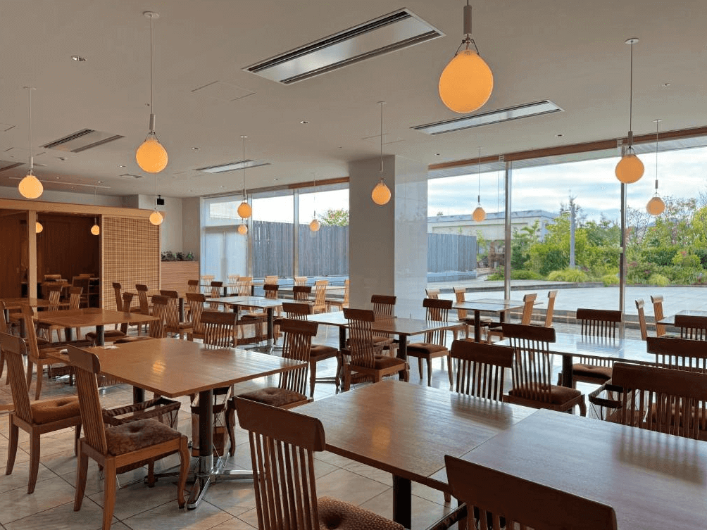【大分住宿】大分JR九州布魯斯姆酒店 (JR Kyushu Hotel Blossom Oita) 的庭園景觀餐廳,室內空間寬敞明亮,懸掛著溫馨的圓形吊燈,排列整齊的木質餐桌椅旁即是落地窗,提供旅客優雅舒適的用餐環境。