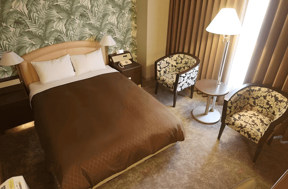 【大分住宿】大分倫勃朗酒店 (Rembrandt Hotel Oita) 的單人客房,牆面採用獨特的綠色熱帶葉片壁紙裝飾,搭配兩張印有古典花卉圖騰的休閒扶手椅與立燈,營造出充滿歐式復古風情的休憩角落。