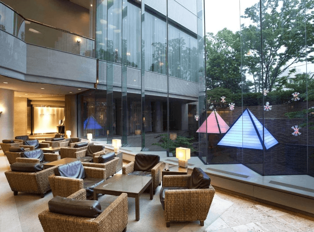 【大分住宿】大分倫勃朗酒店 (Rembrandt Hotel Oita) 的挑高大廳酒吧區域,透過整面落地玻璃窗可欣賞戶外庭園的綠意與幾何金字塔造型燈飾,室內擺設藤編風格的沙發座椅,展現出優雅開闊的度假氛圍。