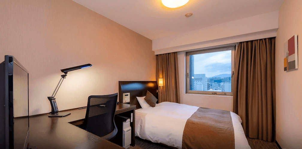 【大分住宿】大分大和ROYNET飯店 (Daiwa Roynet Hotel Oita) 的單人客房,專為商務人士打造,配備寬大的L型轉角辦公桌與符合人體工學的網狀辦公椅,窗外可見市區街景,打造出明亮且高效率的行動辦公室環境。