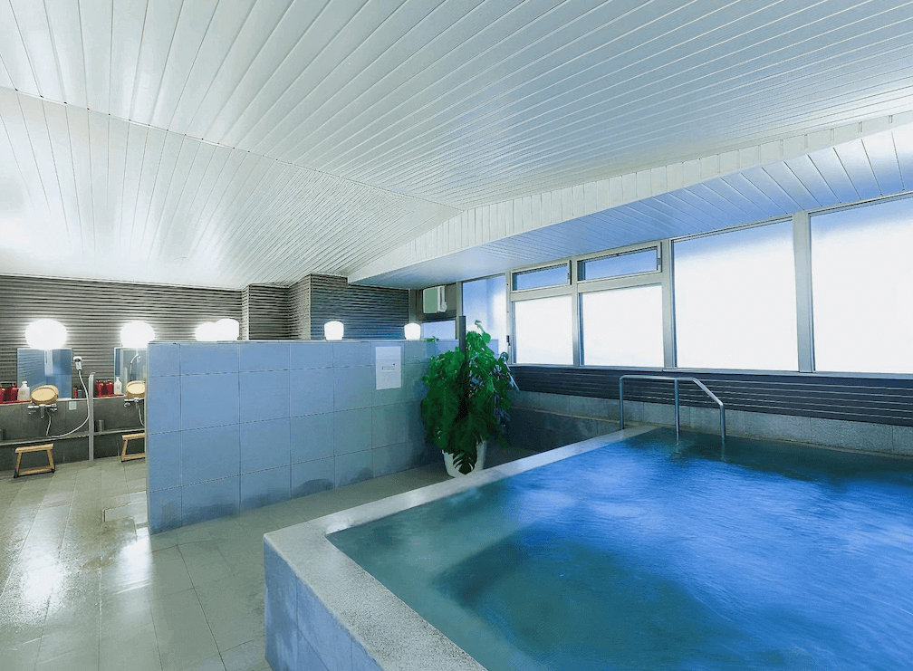 【大分住宿】大分JR九州布魯斯姆酒店 (JR Kyushu Hotel Blossom Oita) 頂樓「CITY SPA 天空」的室內大浴場,擁有寬敞的石造浴池與溫暖的木質窗框,透過整面落地大窗,旅客可在泡湯的同時飽覽大分市區的壯闊景致。