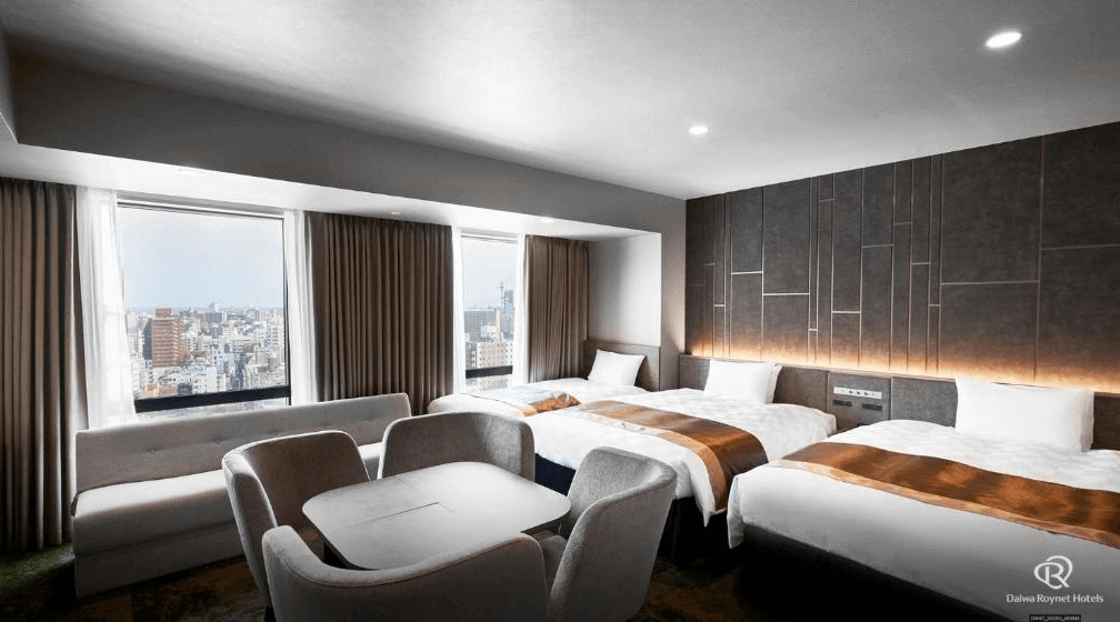 【鹿兒島住宿】大和魯內酒店鹿兒島天文館PREMIER (Daiwa Roynet Hotel Kagoshima Tenmonkan PREMIER) 的寬敞家庭房型，設有三張舒適單人床，並規劃出包含L型沙發與圓桌的獨立起居空間，窗外即可俯瞰鹿兒島市區街景。