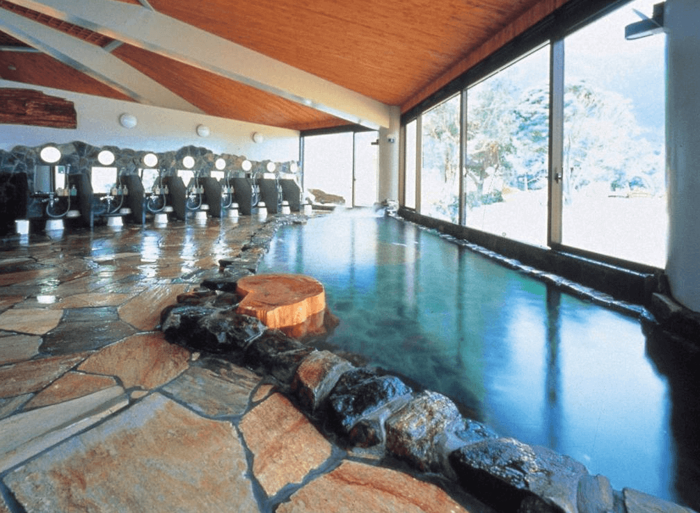 【屋久島住宿】「屋久島岩崎飯店（Yakushima Iwasaki Hotel）溫泉大浴場，以岩石與木質元素打造自然風格，落地窗可望向山林景色。」