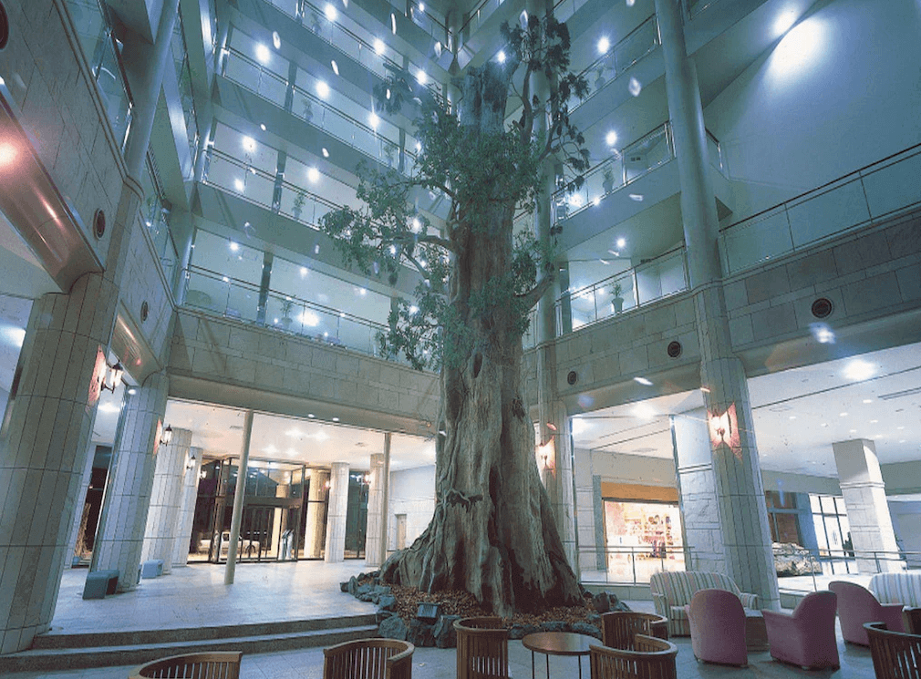 【屋久島住宿】「屋久島岩崎飯店（Yakushima Iwasaki Hotel）挑高大廳中央矗立象徵屋久杉的巨大裝飾樹木，周圍環繞著座位區與飯店樓層。」