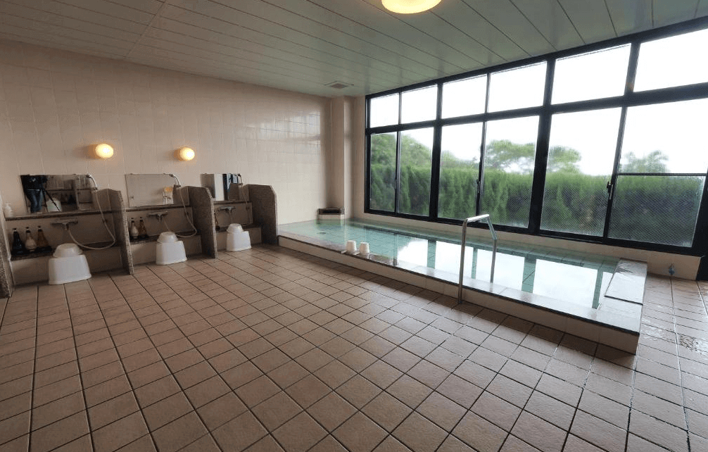 【屋久島住宿】「屋久島綠色酒店室內大浴場，設有多個梳洗座位與大型溫泉池，大片窗景可欣賞戶外綠意，提供旅客放鬆泡湯的舒適環境。」