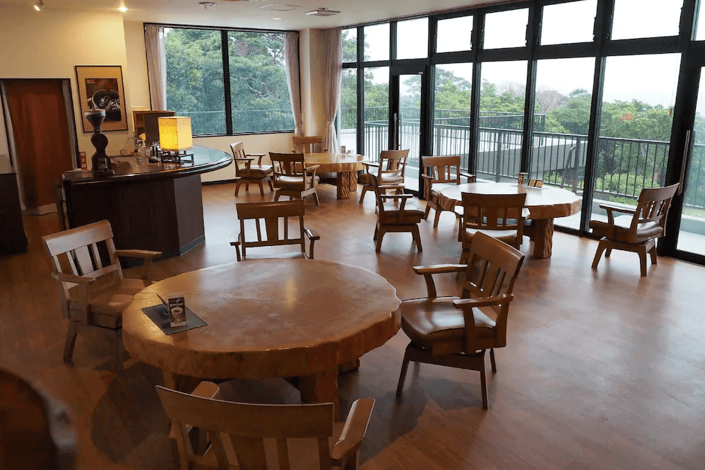【屋久島住宿】「屋久島綠色酒店館內咖啡廳，使用大面積玻璃窗引入自然光，木質桌椅與寬敞座位區營造溫暖放鬆的氛囲，可享受悠閒飲品時光。」