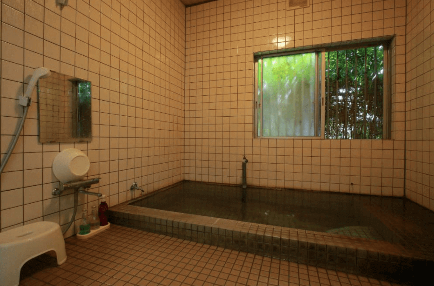 【指宿住宿】川久家庭旅館 (Family Ryokan Kawakyu) 的室內浴場，採用復古的白色磁磚設計，設有寬敞的長方形浴池，窗外可見翠綠的竹林景致，營造出寧靜的洗浴氛圍。