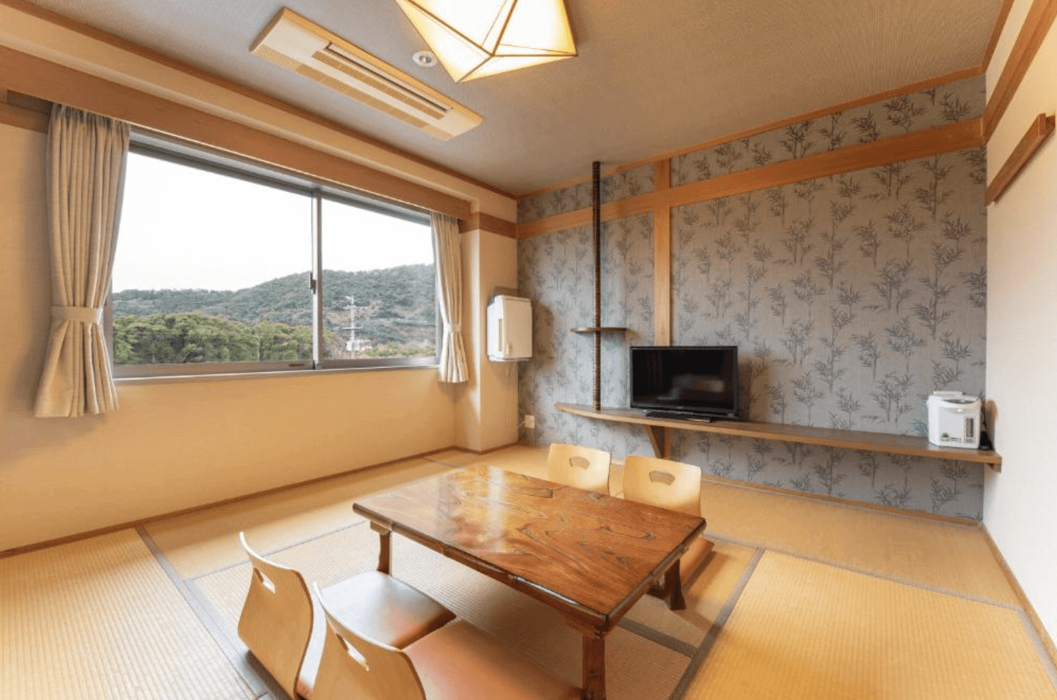【指宿住宿】指宿Kokoro之宿 (Hotel Ibusuki Kokoronoyado) 的日式和室客房，鋪設傳統榻榻米與原木矮桌，牆面裝飾著雅緻的圖樣，窗外是一片綠意盎然的山林景色，適合喜愛寧靜的旅客。