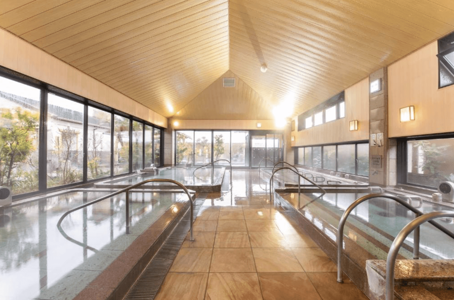 【指宿住宿】指宿Kokoro之宿 (Hotel Ibusuki Kokoronoyado) 的大浴場，擁有挑高的木造斜屋頂設計與寬敞的室內空間，透過大面落地窗引入自然光線，讓旅客在開闊的環境中享受舒適的溫泉。