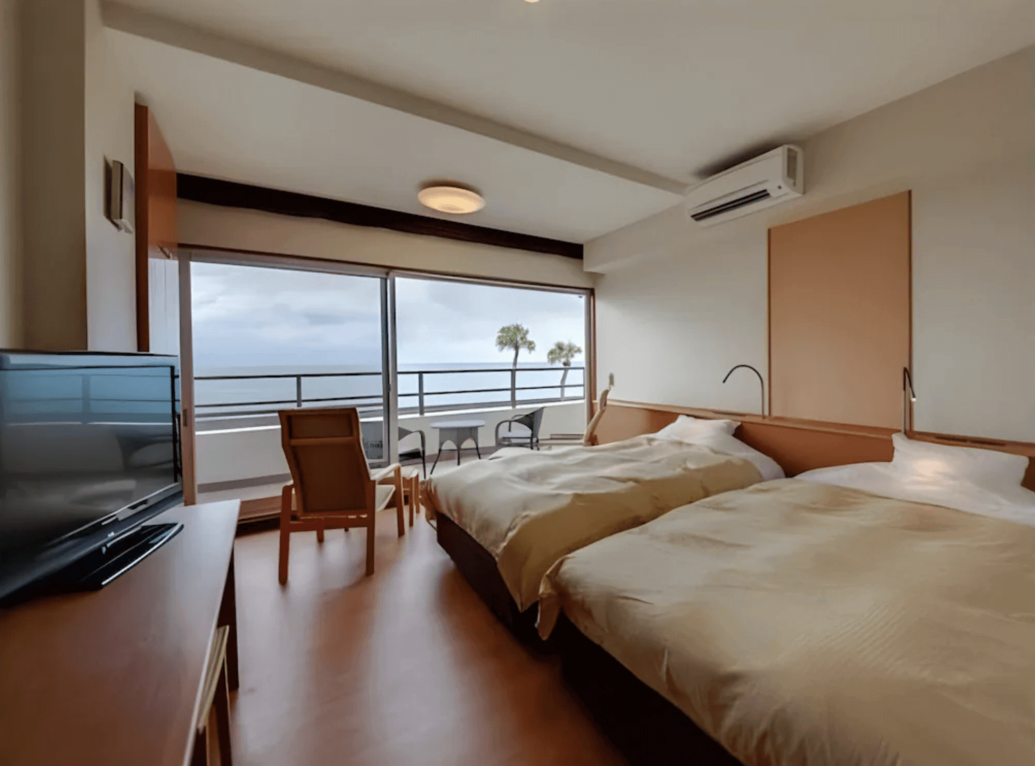 【指宿住宿】指宿休暇村 (Kyukamura Ibusuki) 的洋式雙床客房，設計簡約明亮，落地窗外設有專屬觀景陽台，旅客躺在床上即可欣賞窗外隨風搖曳的棕櫚樹與蔚藍海平線，充滿南國度假氣息。