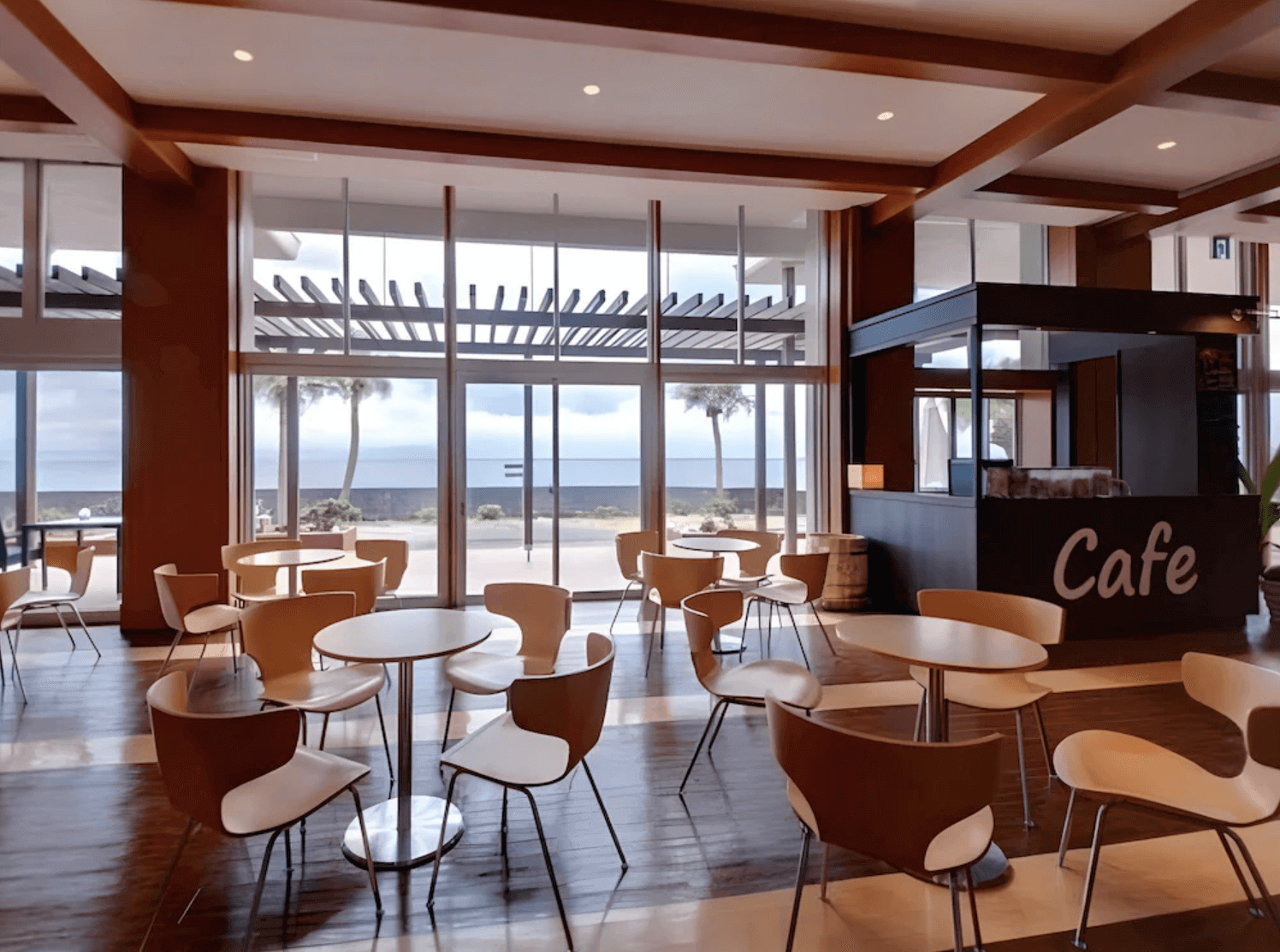 【指宿住宿】指宿休暇村 (Kyukamura Ibusuki) 的開放式大廳咖啡座區域，擁有挑高的木造天花板與通透的落地玻璃窗，陽光灑落在設計感十足的木質桌椅上，窗外即是寧靜的海岸風光。